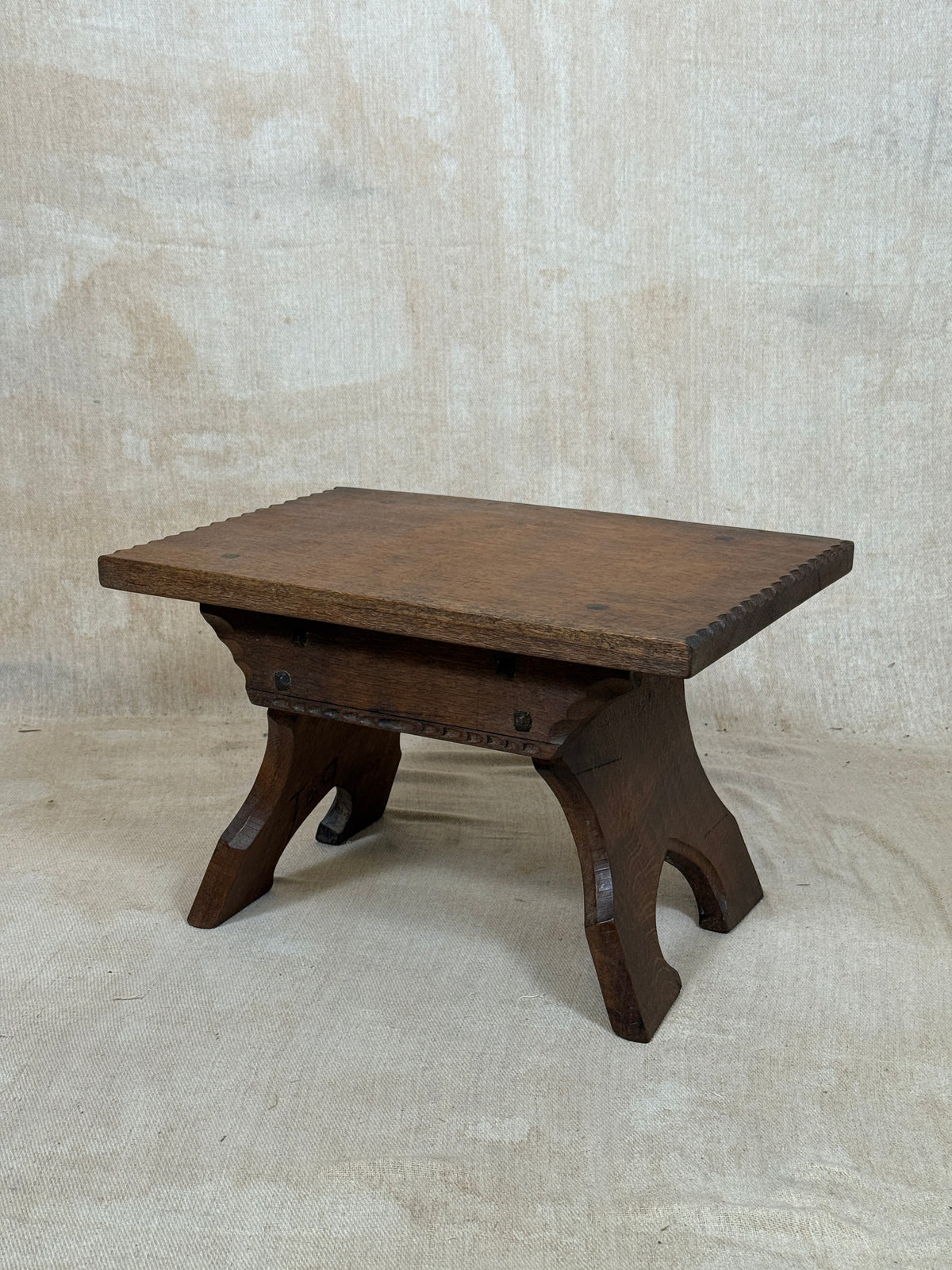 A dark brown wooden stool with a rectangular top and curved legs, photographed against a textured beige fabric backdrop.