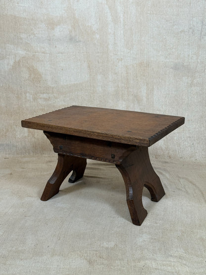 A dark brown wooden stool with a rectangular top and curved legs, photographed against a textured beige fabric backdrop.
