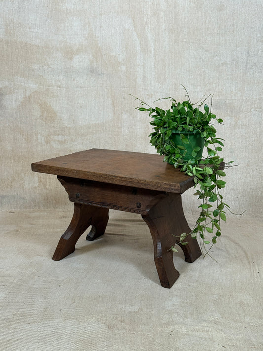 A rustic dark brown wooden stool with a rectangular top and three curved legs, styled with a green trailing plant on a textured beige fabric backdrop.