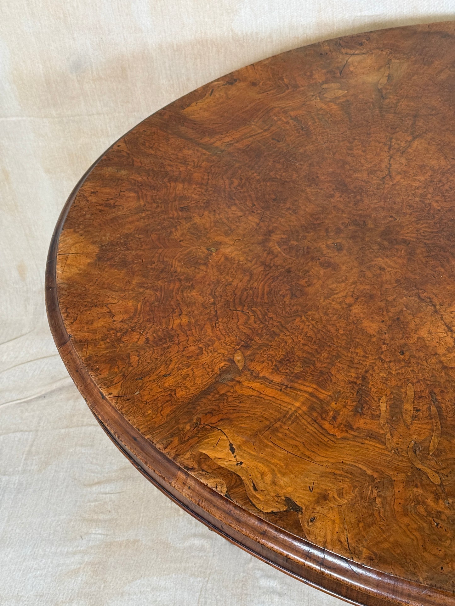 Close-up of a polished oval wooden tabletop with dramatic burl wood grain in rich brown tones, set against a light beige fabric background.