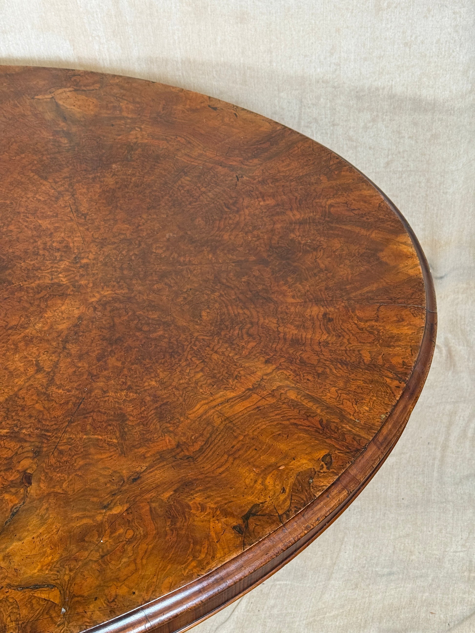 Close-up of a polished burl wood tabletop showing rich brown grain patterns and a curved edge, set against a light fabric background.