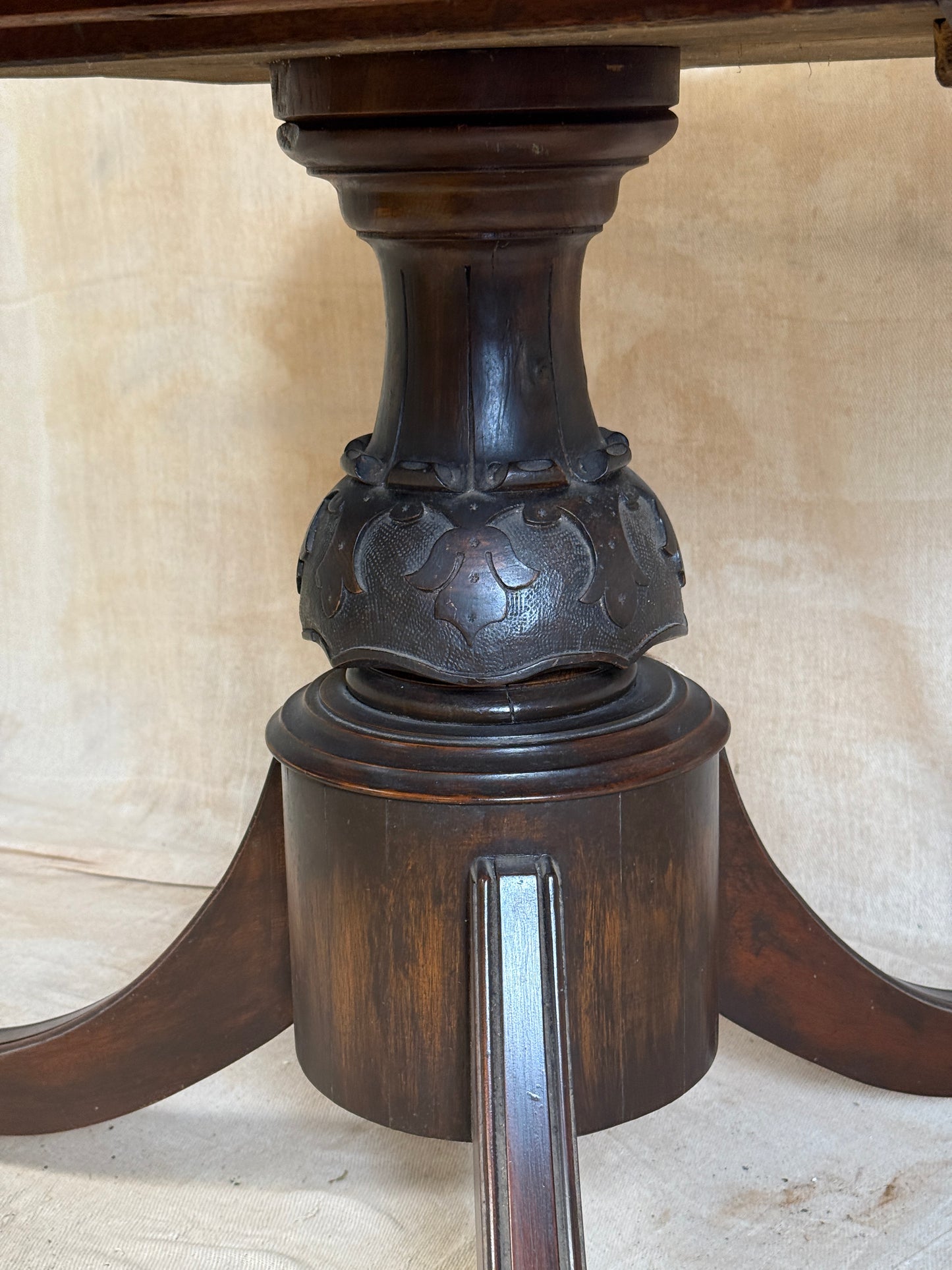 Close-up of a dark brown carved wooden table pedestal base with a metal bar extending from the side, set against a light beige tiled background.
