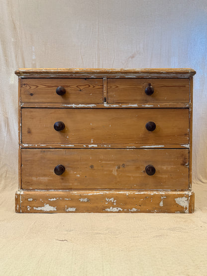 A rustic pine wooden chest of drawers with three drawers, dark round knobs, and distressed white paint showing through the weathered finish, set against a neutral fabric backdrop.