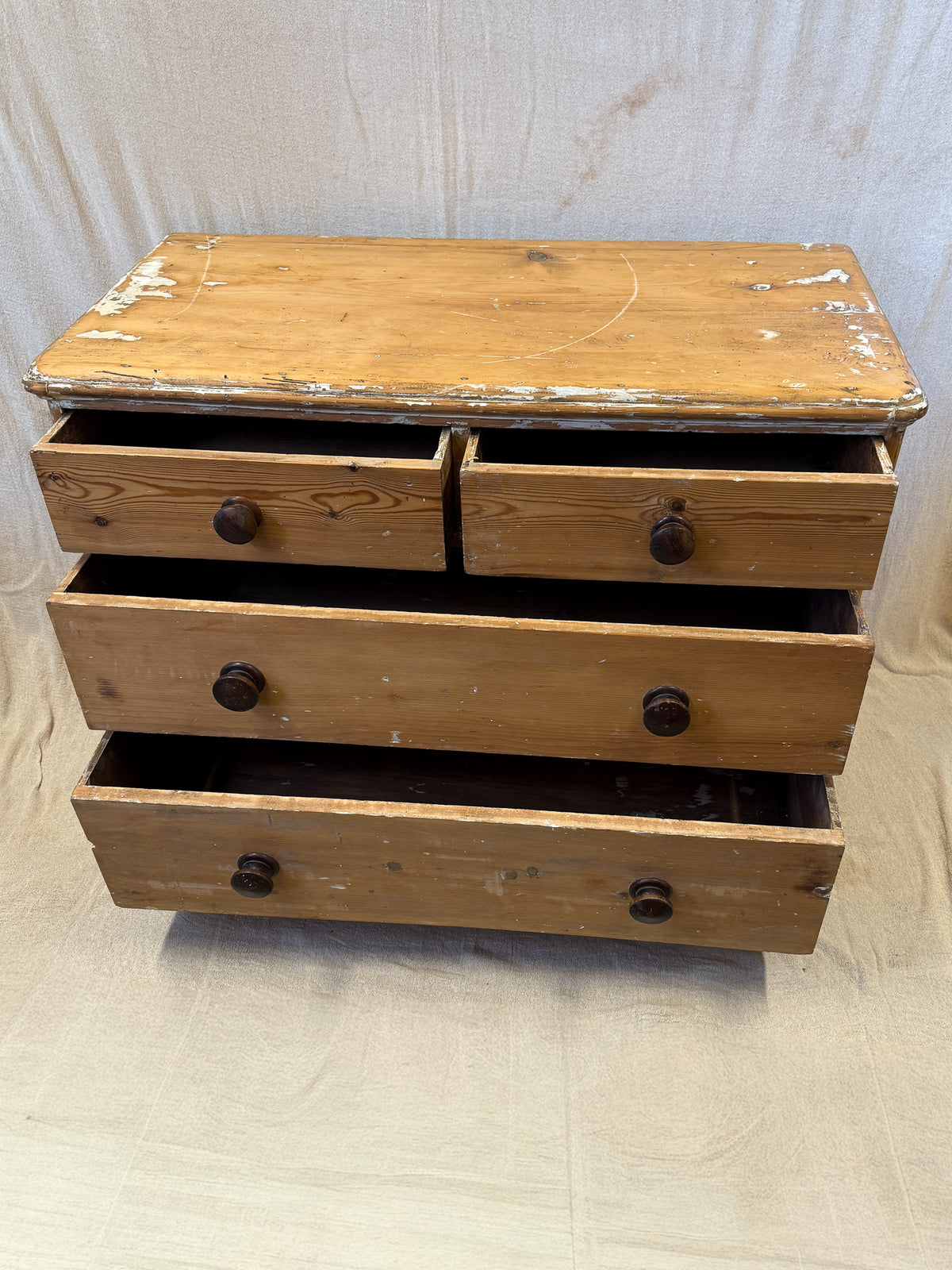 A rustic wooden chest of drawers with a distressed finish, featuring a top surface and three drawers with dark round knobs, set against a light fabric backdrop.
