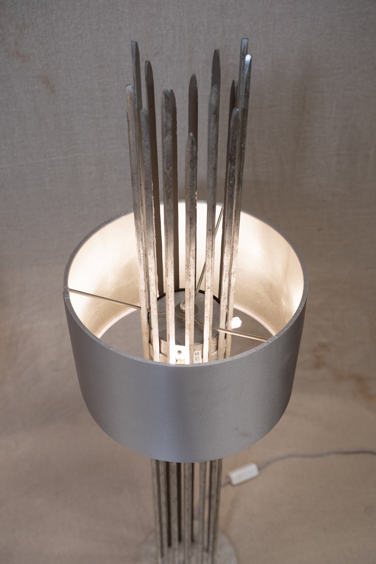 A modern sculptural floor lamp with a wide cylindrical silver shade and multiple vertical metallic rods, lit from within against a neutral fabric backdrop.