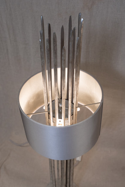 A modern sculptural floor lamp featuring a wide, matte silver cylindrical shade with multiple slender metallic rods rising from the center, set against a neutral draped fabric backdrop.