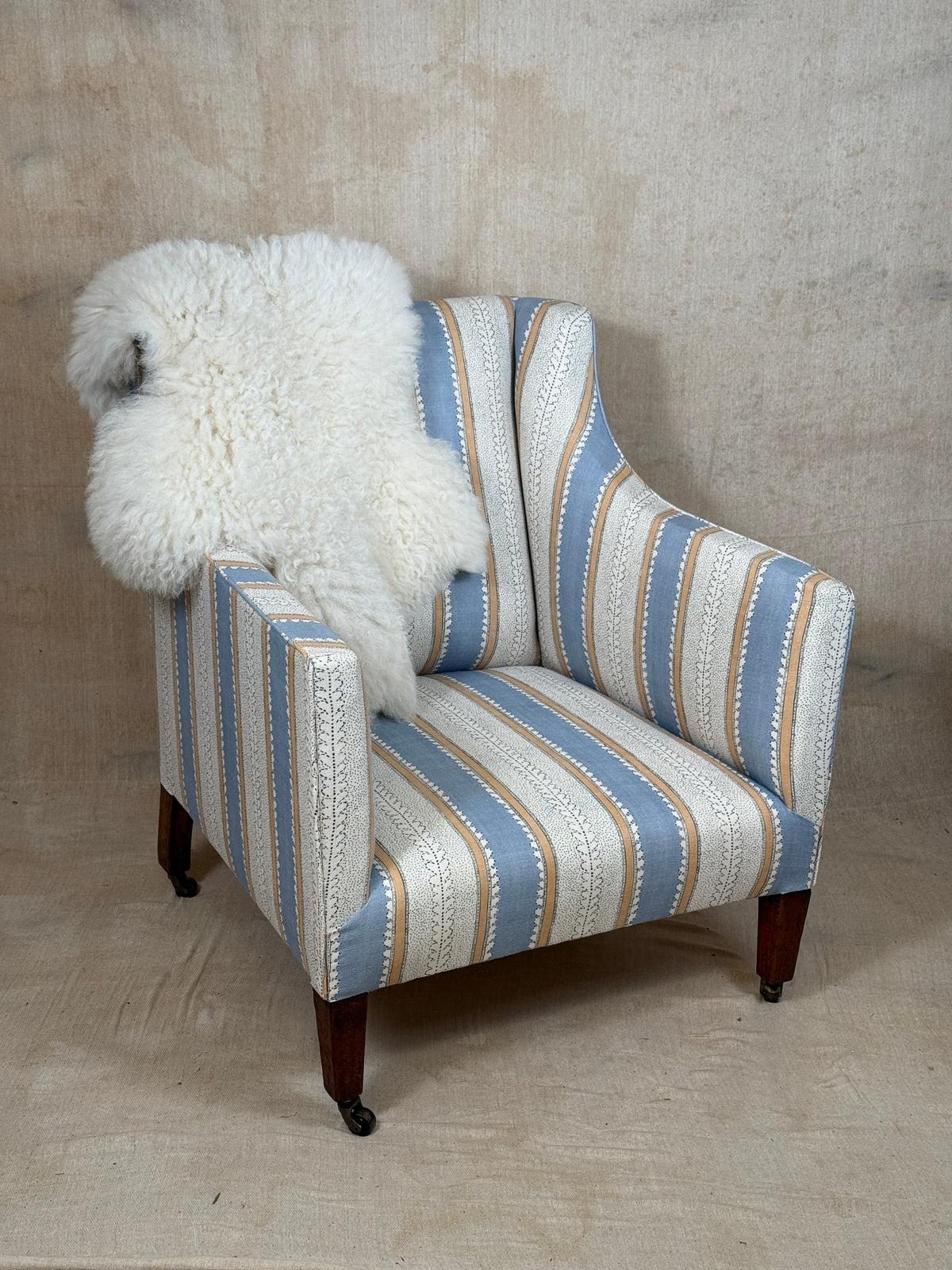 A vintage-style wingback armchair with blue, beige, and white striped upholstery, featuring dark wood legs and a fluffy white sheepskin throw draped over the back, set against a textured beige backdrop.