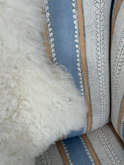 Close-up of a textured white sheepskin throw pillow resting on a chair upholstered in a light blue and white patterned fabric with beige stripes.