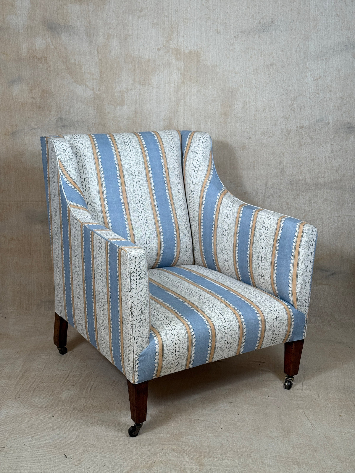 A wingback armchair upholstered in a vertical striped fabric with light blue, cream, and tan stripes, featuring dark wood legs with black casters, set against a textured concrete wall and floor.