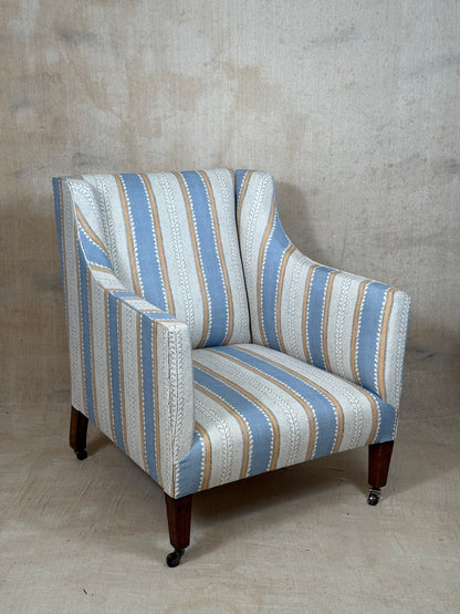 A wingback armchair upholstered in a vertical striped fabric with light blue, cream, and tan stripes, featuring dark wood legs with black casters, set against a textured concrete wall and floor.
