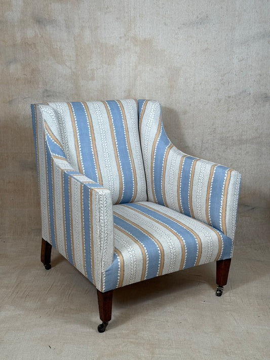 A wingback armchair upholstered in a vertical striped fabric with light blue, cream, and tan stripes, featuring dark wood legs with black casters, set against a textured concrete wall and floor.