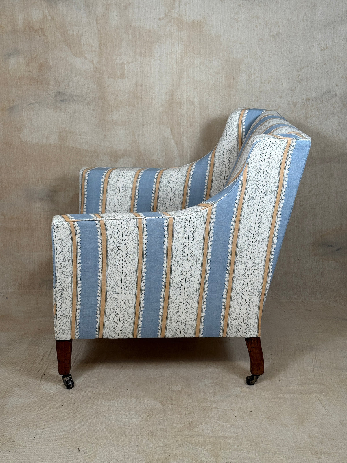 A single upholstered armchair shown from the side against a textured beige studio backdrop. The chair has a striped fabric pattern with vertical bands of light blue, off-white, tan, and cream, plus small white geometric motifs.