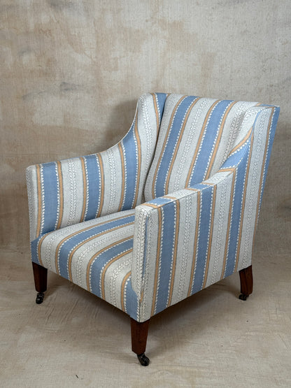 A wingback armchair upholstered in a vertical striped fabric with blue, beige, and white tones, featuring dark wood legs with casters, set against a textured concrete wall.