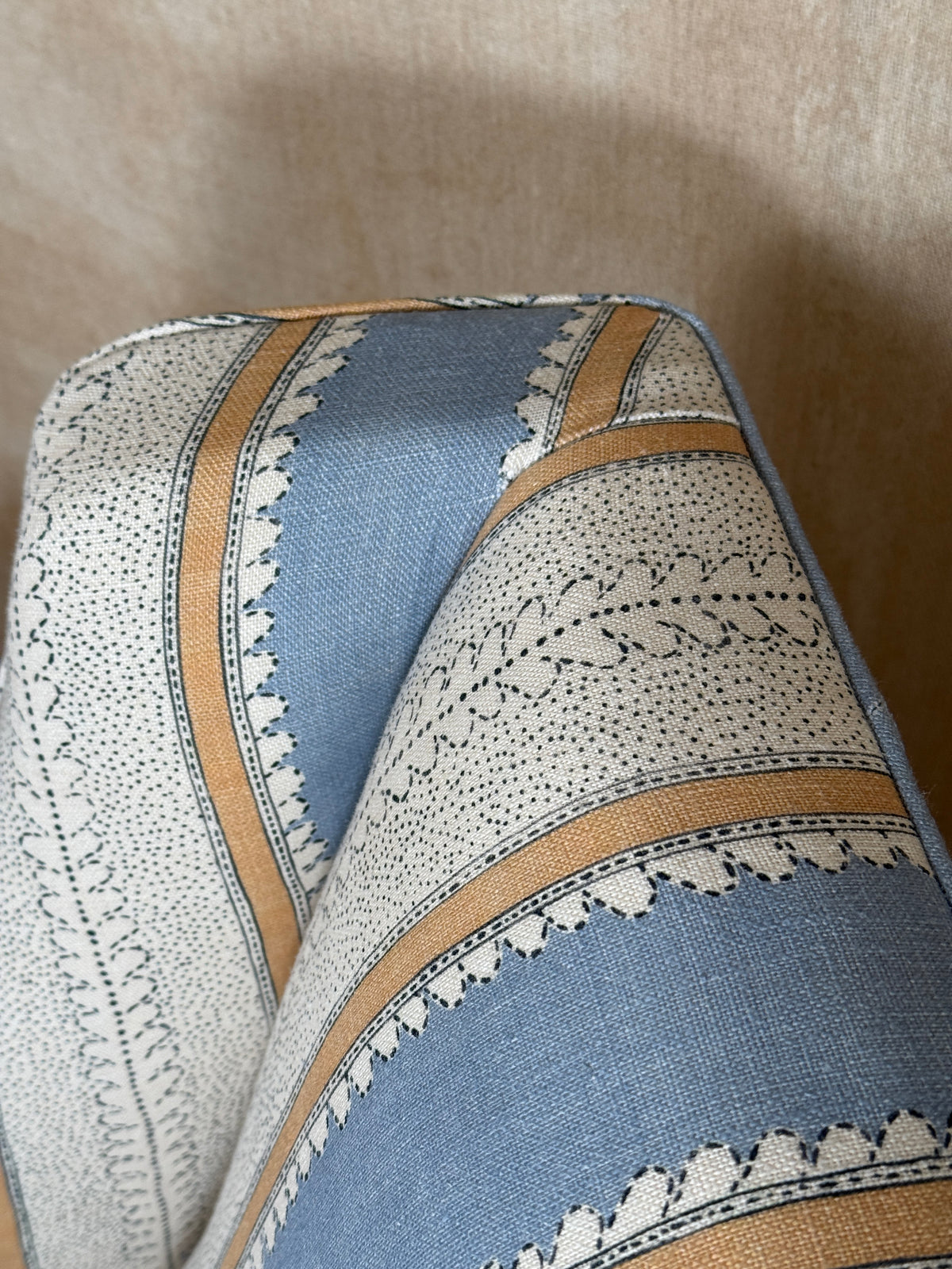 A close-up of patterned upholstery fabric with alternating blue and off-white sections, decorated with black leaf motifs and bordered by tan stripes, set against a beige background.
