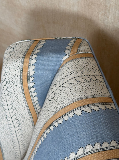 A close-up of patterned upholstery fabric with alternating blue and off-white sections, decorated with black leaf motifs and bordered by tan stripes, set against a beige background.