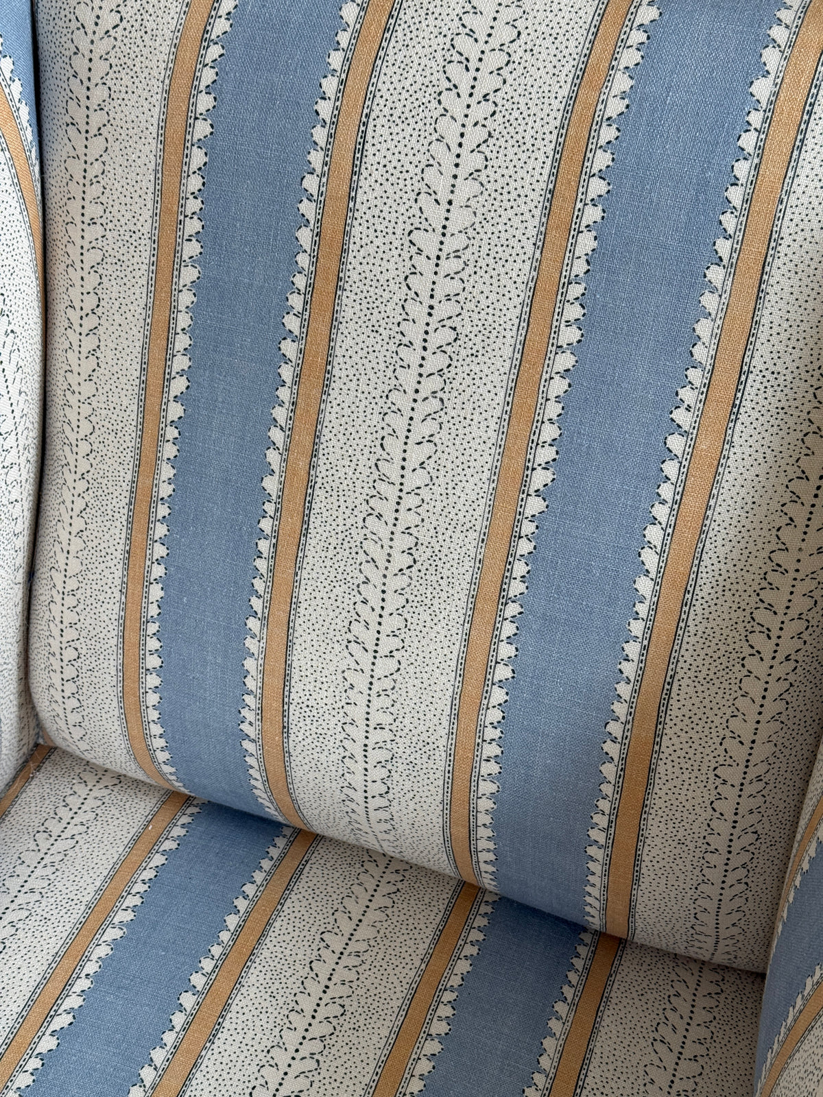 Close-up of upholstered fabric with vertical stripes in dusty blue, warm tan, and off-white with small leafy lace-like motifs.