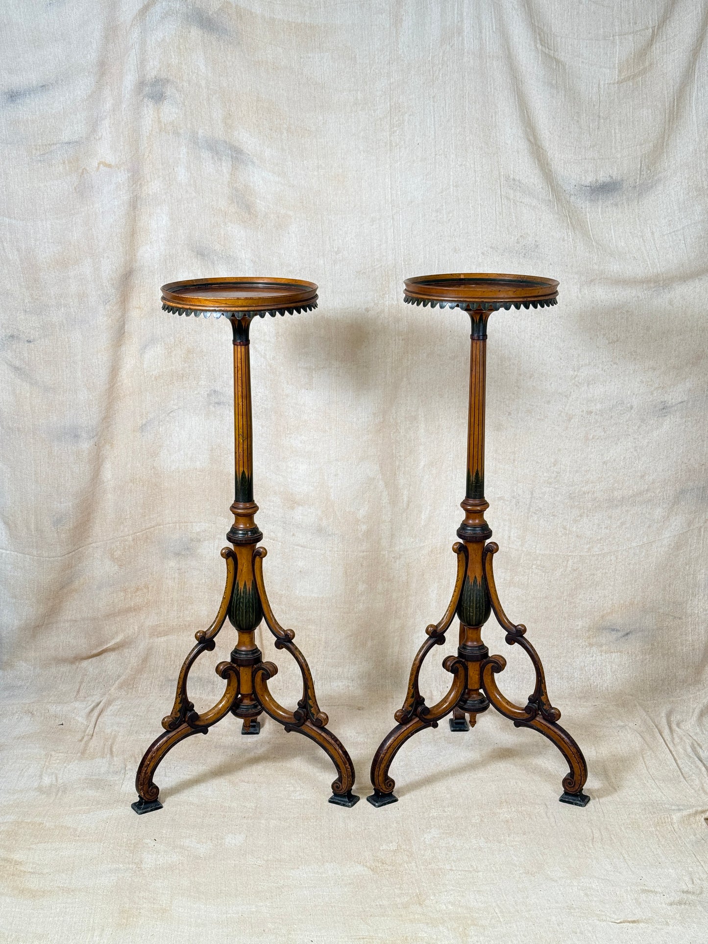 Two ornate dark brown Torchere Stands with decorative scrollwork bases, shown against a draped beige fabric backdrop. 