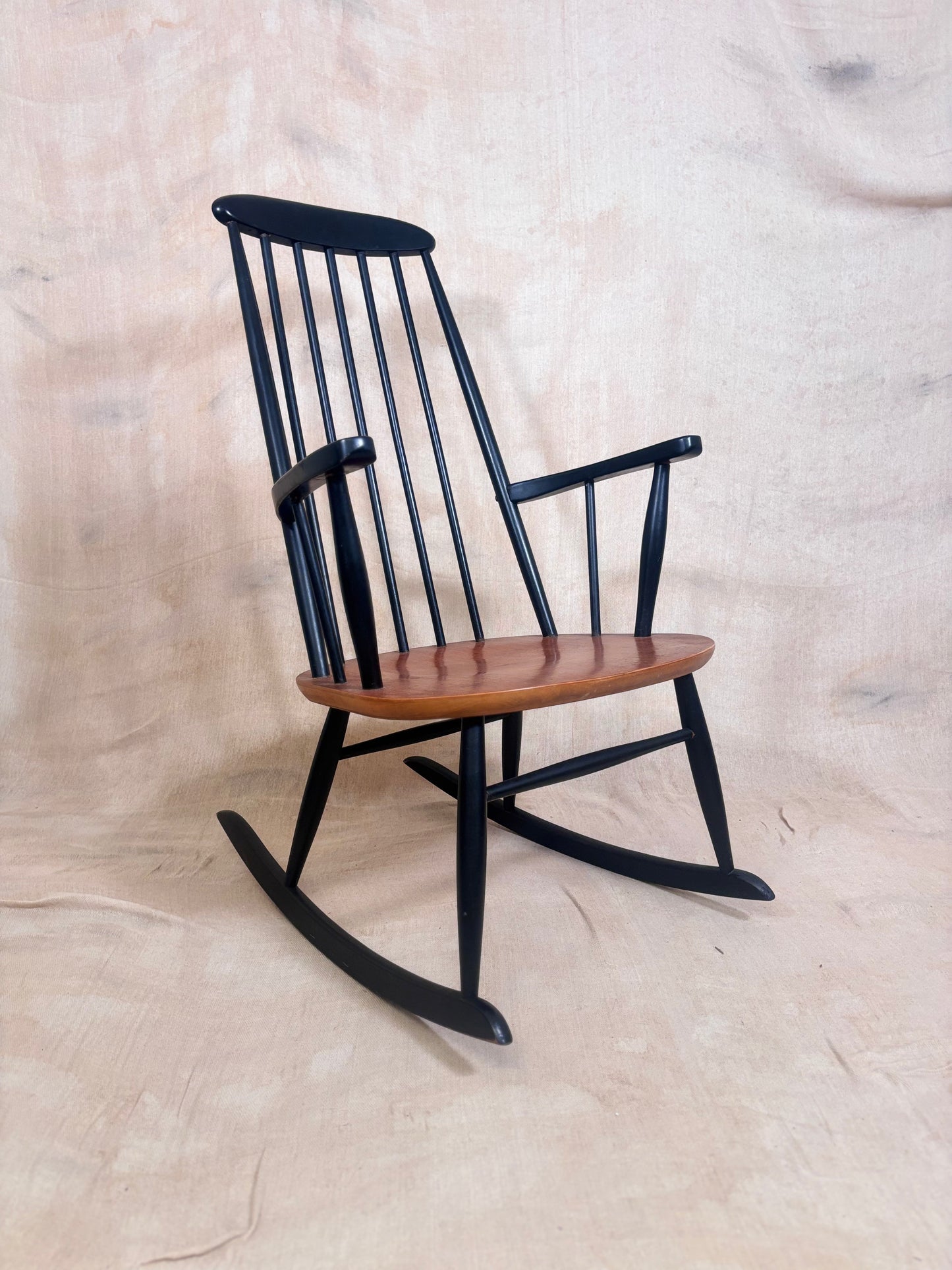 A classic wooden rocking chair with a black spindle-back frame and a warm brown wooden seat, photographed against a neutral beige fabric backdrop.
