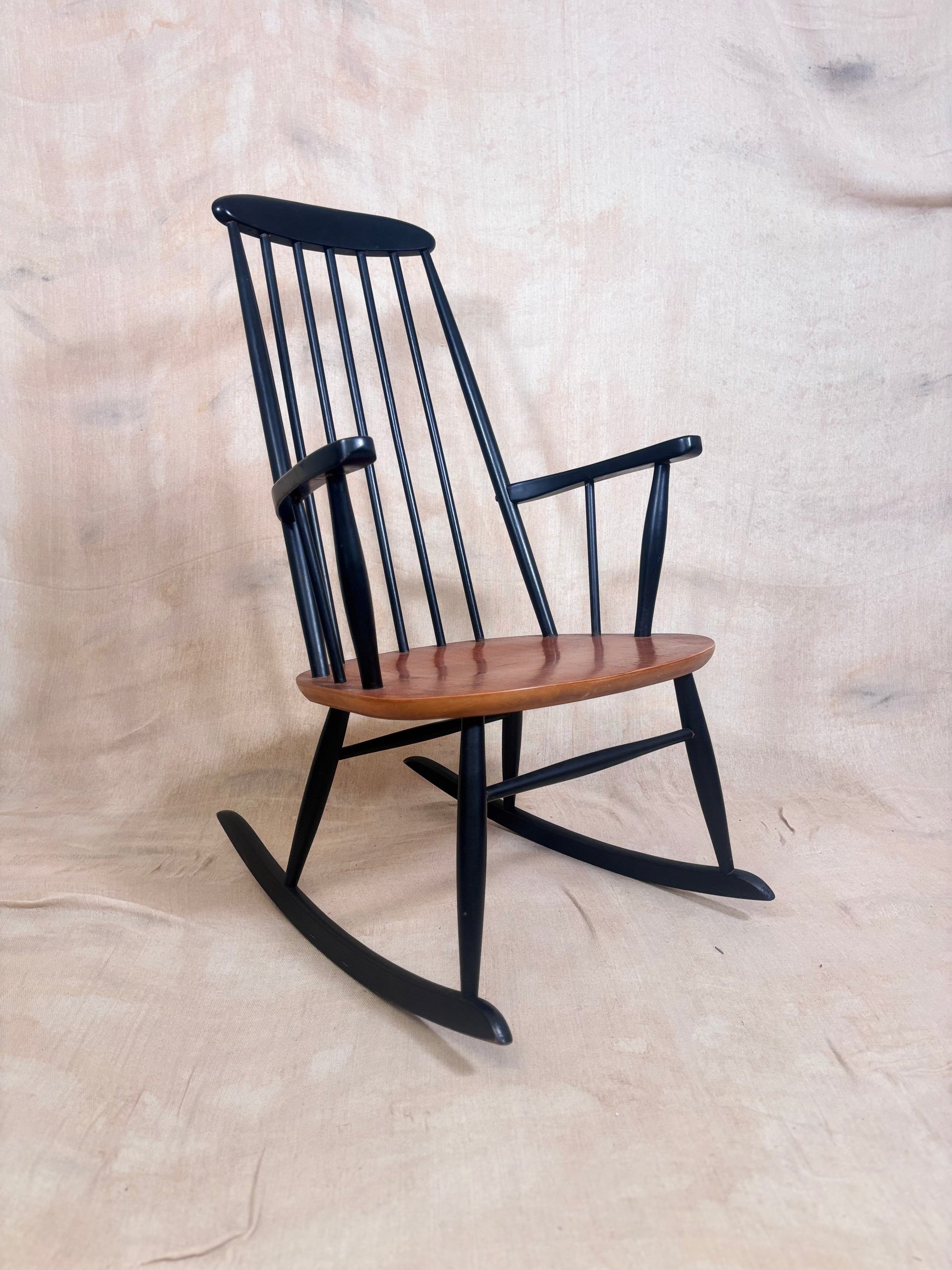 A classic wooden rocking chair with a black spindle-back frame and a warm brown wooden seat, photographed against a neutral beige fabric backdrop.