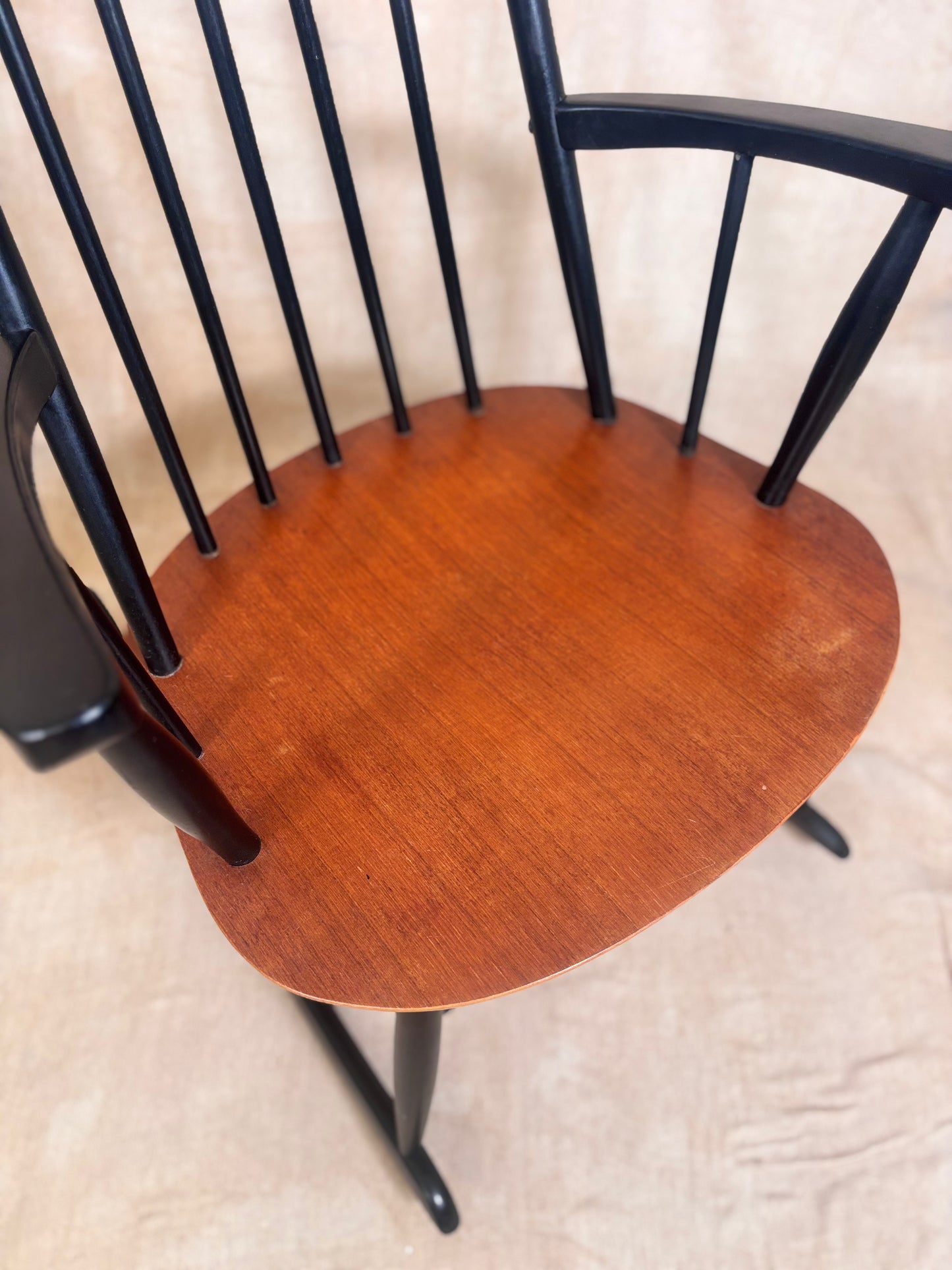 A close-up of a wooden chair with a warm reddish-brown spindle back and black painted frame, shown against a light beige textured background.
