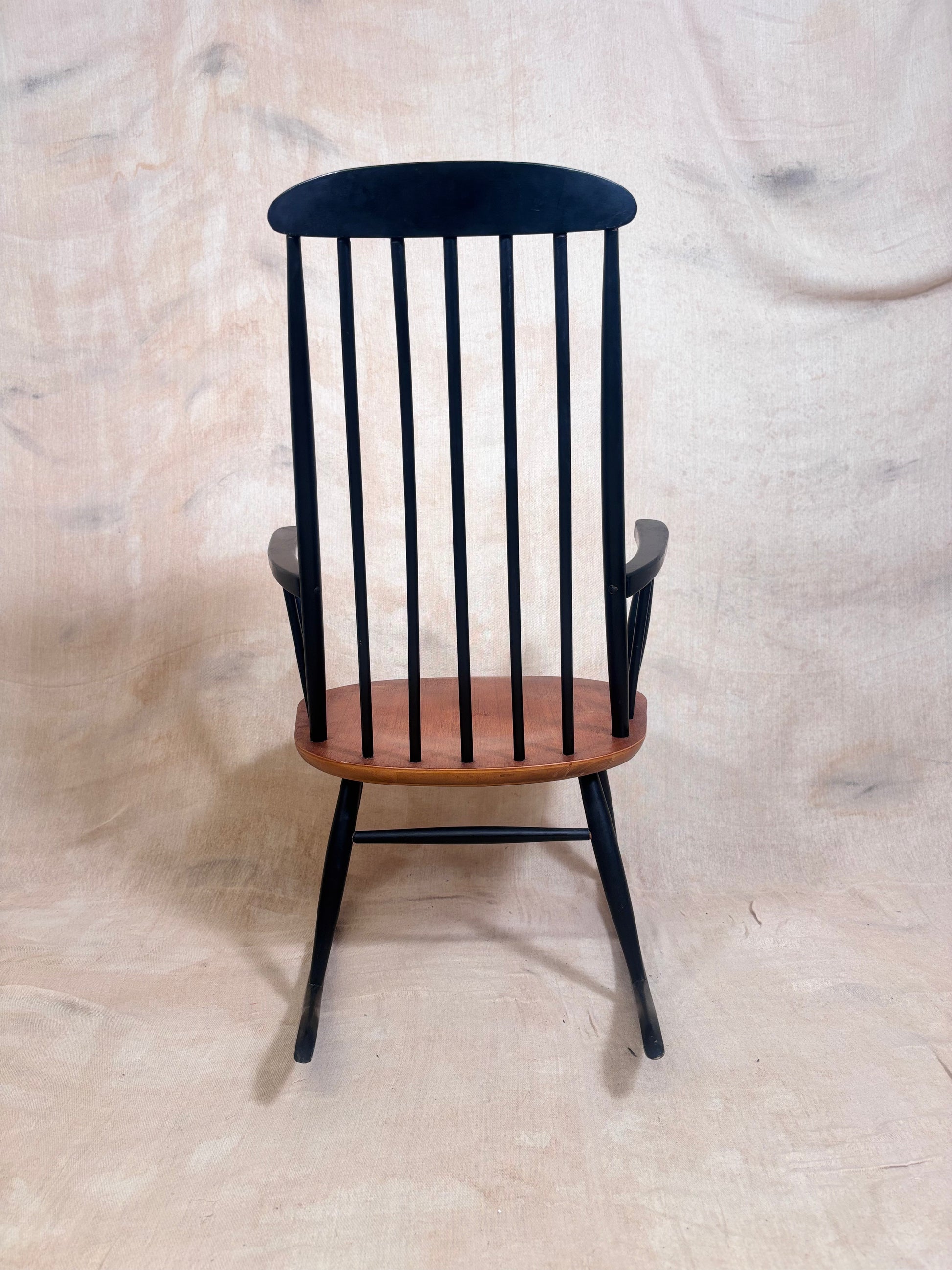 A black spindle-back rocking chair with a warm brown wooden seat, photographed against a draped beige fabric backdrop.