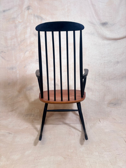 A black spindle-back rocking chair with a warm brown wooden seat, photographed against a draped beige fabric backdrop.