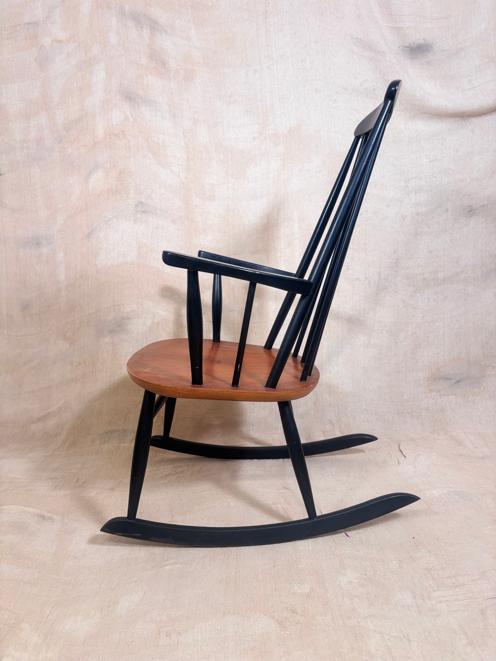 A classic wooden rocking chair with a black spindle-back frame and a warm brown wooden seat, photographed against a neutral beige fabric backdrop.