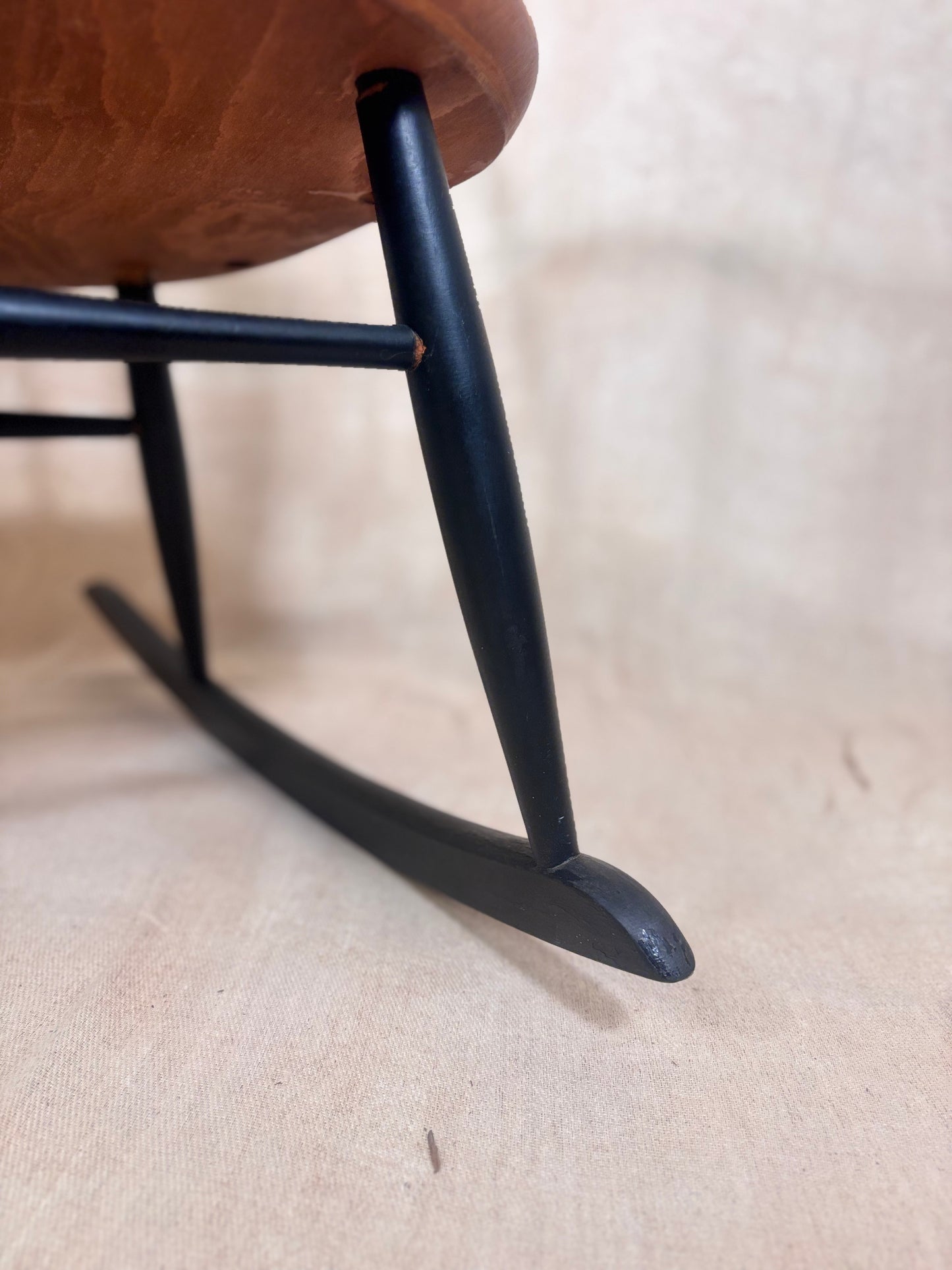 Close-up of a rocking chair's black painted wooden legs and curved rockers, with part of the warm brown upholstered seat visible against a light beige fabric background.