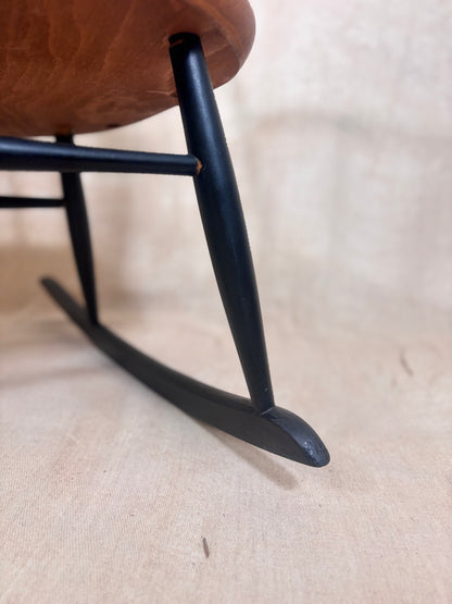 Close-up of a rocking chair's black painted wooden legs and curved rockers, with part of the warm brown upholstered seat visible against a light beige fabric background.