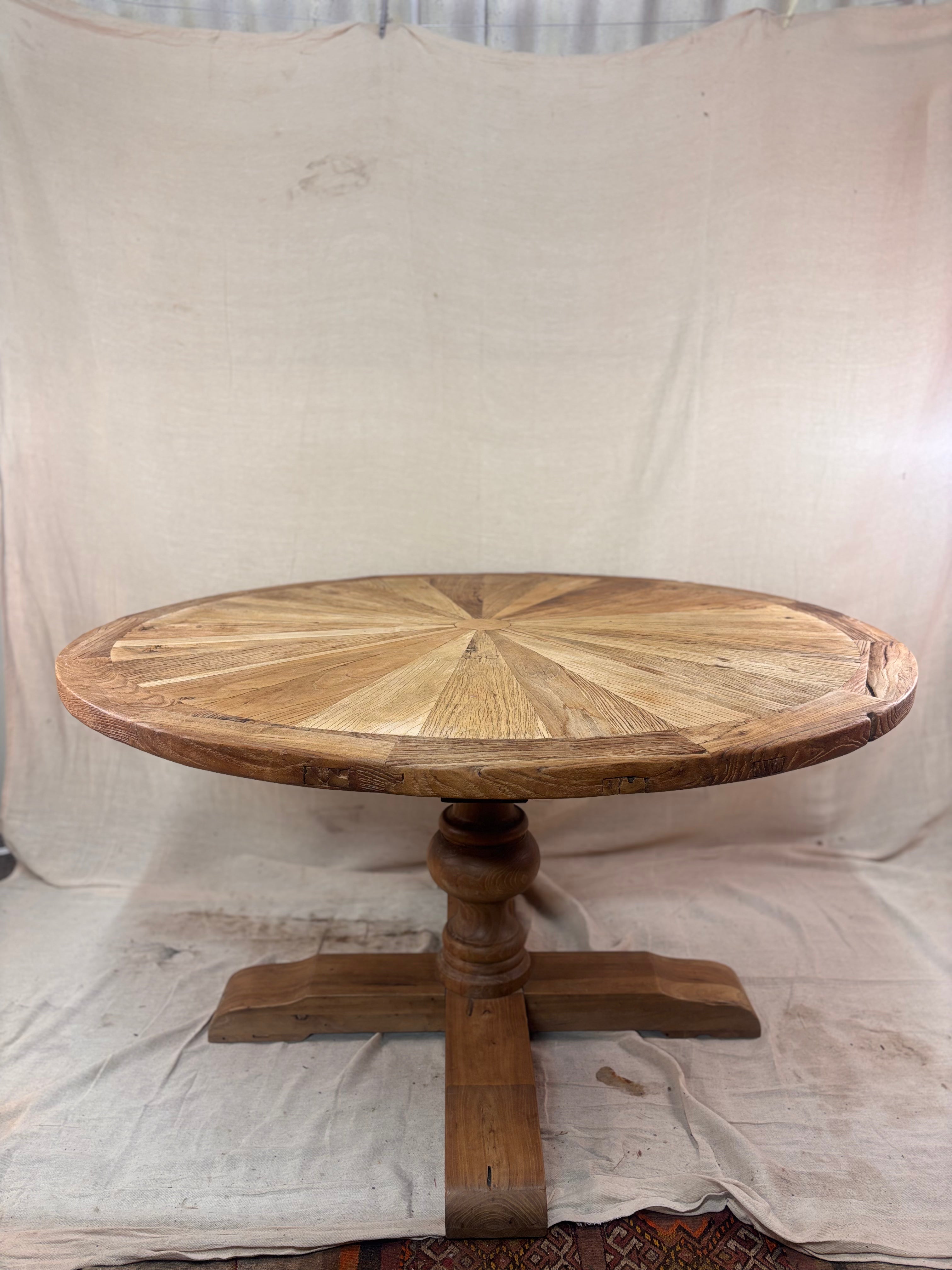 Rustic round wooden pedestal dining table with sunburst-patterned top and cross-leg base, photographed against a neutral backdrop.