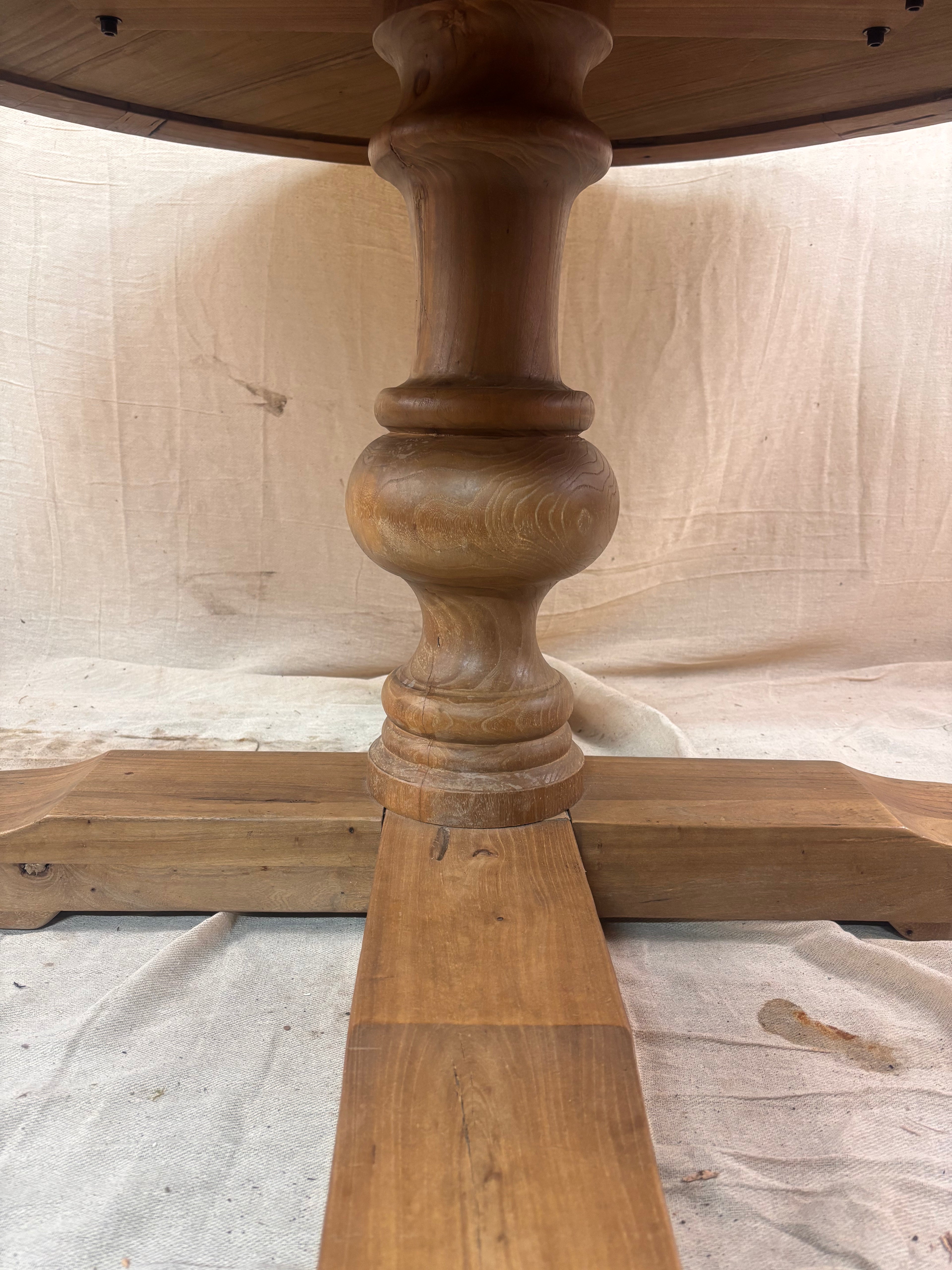 Rustic round wooden pedestal dining table with sunburst-patterned top and cross-leg base, photographed against a neutral backdrop.