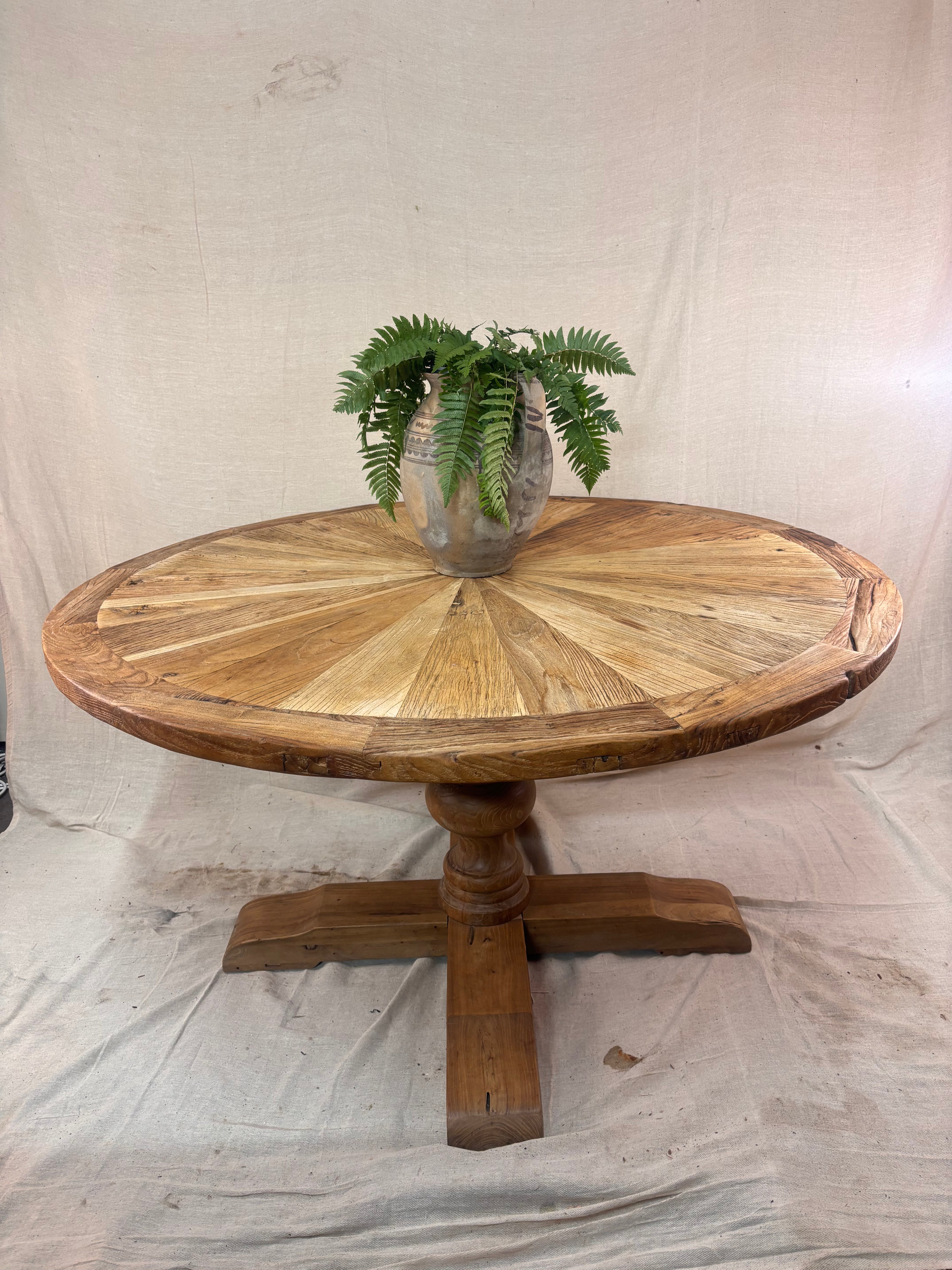 Rustic round wooden pedestal dining table with sunburst-patterned top and cross-leg base, photographed against a neutral backdrop.