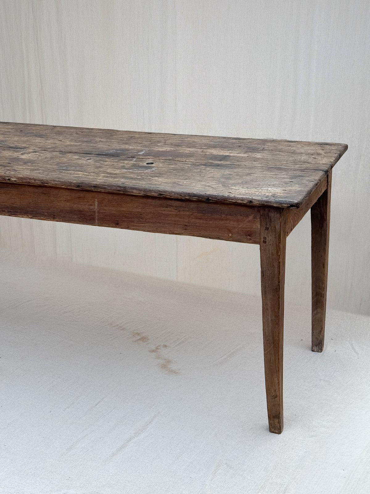 A rustic dark brown wooden dining table with a weathered, distressed top and tapered legs, photographed against a light off-white fabric backdrop.