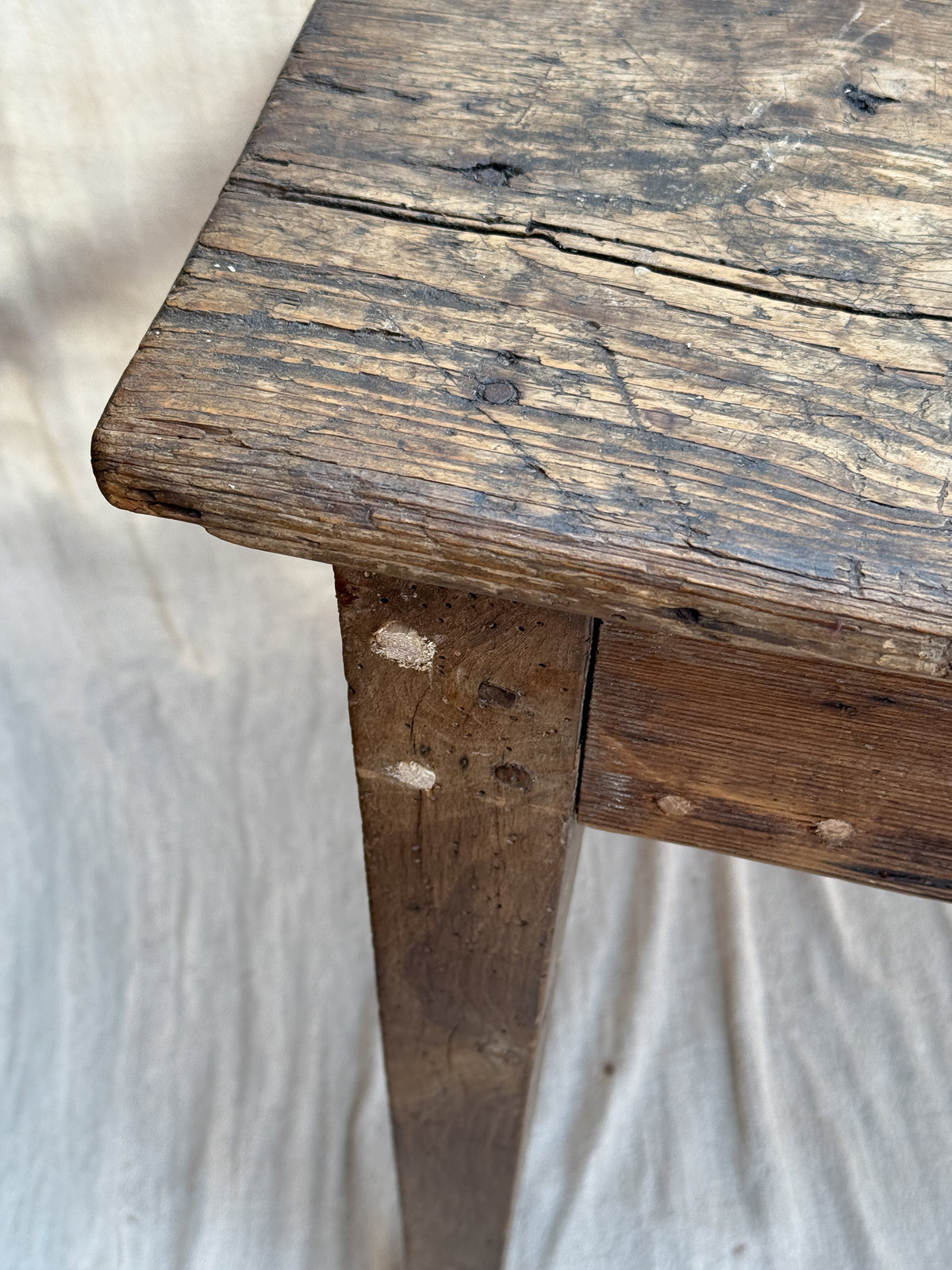 Close-up of a rustic dark brown wooden table corner showing the weathered wood grain, cracks, and natural finish, set against a light fabric background.