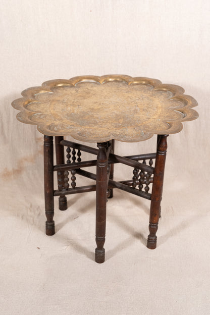 An antique round side table featuring a scalloped brass top and a dark brown wooden base with decorative stretchers, set against a neutral off-white fabric backdrop.