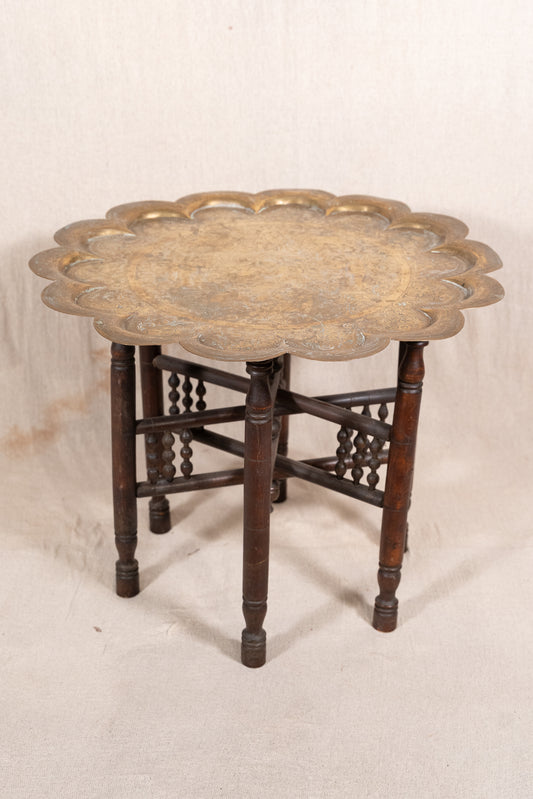 An antique round side table featuring a scalloped brass top and a dark brown wooden base with decorative stretchers, set against a neutral off-white fabric backdrop.