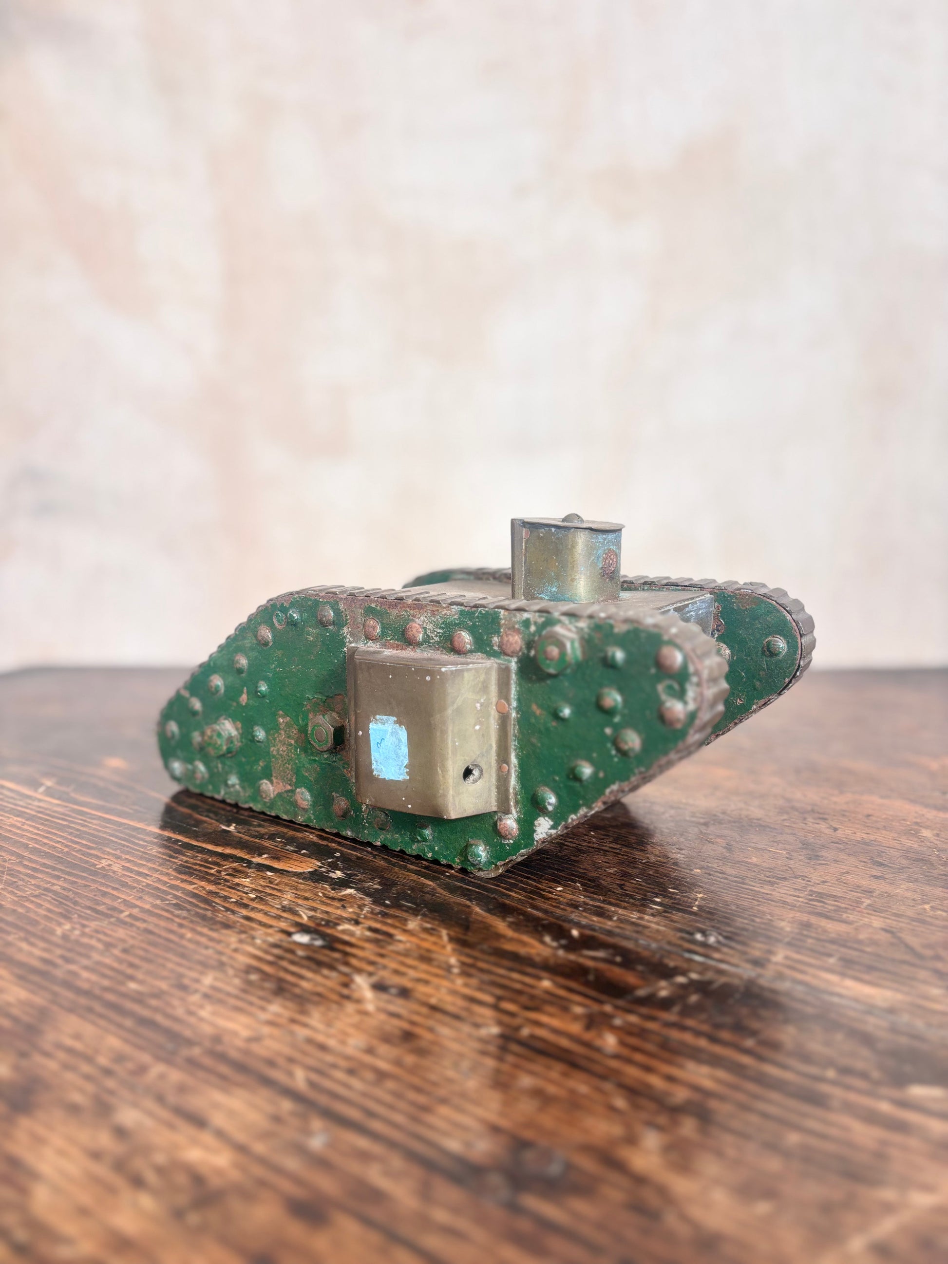 A vintage green metal tank toy or model with rivets and a small blue accent, resting on a rustic wooden table against a textured beige background.