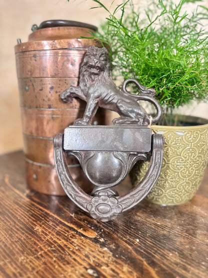 A vintage-style cast iron lion door knocker with a large ring handle, displayed on a rustic wooden table next to a copper bucket and a potted green plant.