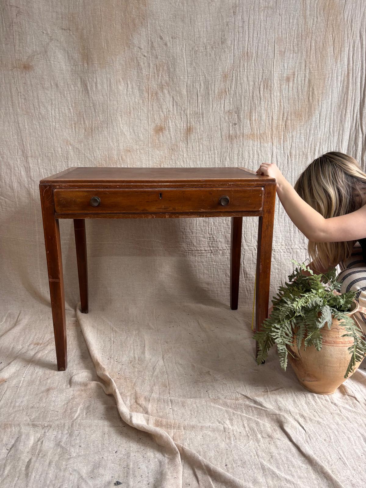 Wooden desk with a drawer against a textured beige wall, with a person and plant in the background.
