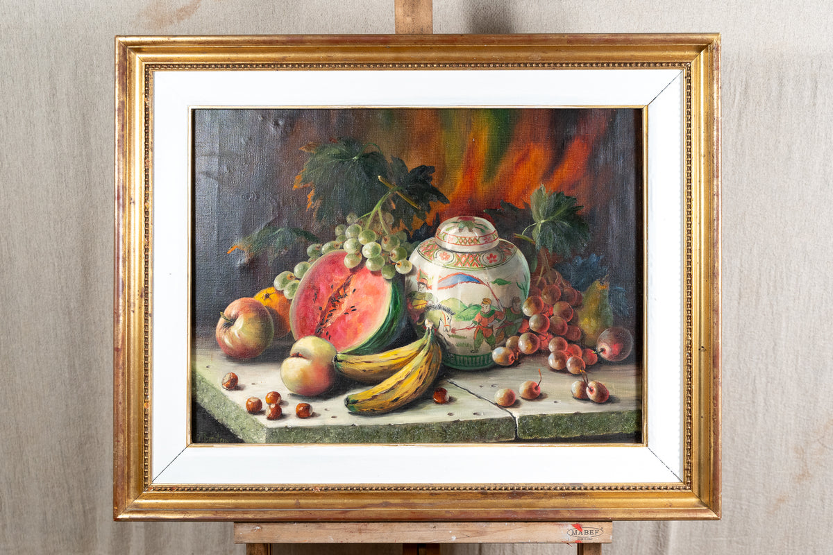 A framed still life oil painting depicting a watermelon, grapes, bananas, apples, and a decorative ceramic jar on a tabletop, displayed on an easel against a neutral wall.