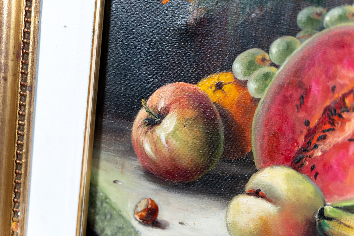 Close-up of a framed still life painting showing fruit, including a red apple, green grapes, a pink watermelon slice, and a hazelnut, with part of the ornate gold frame visible on the left.