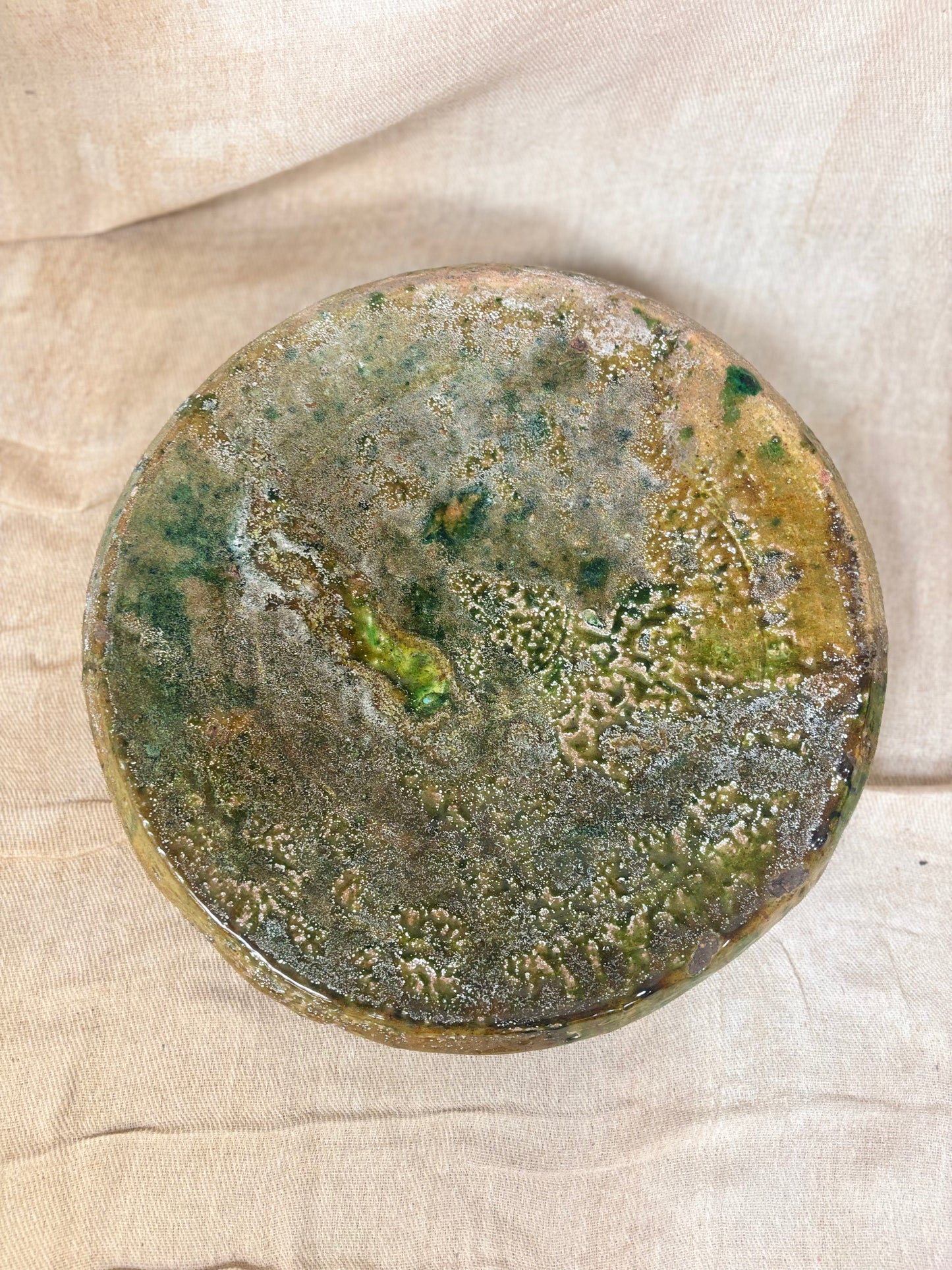 Round ceramic plate with green and brown glaze on a beige fabric background