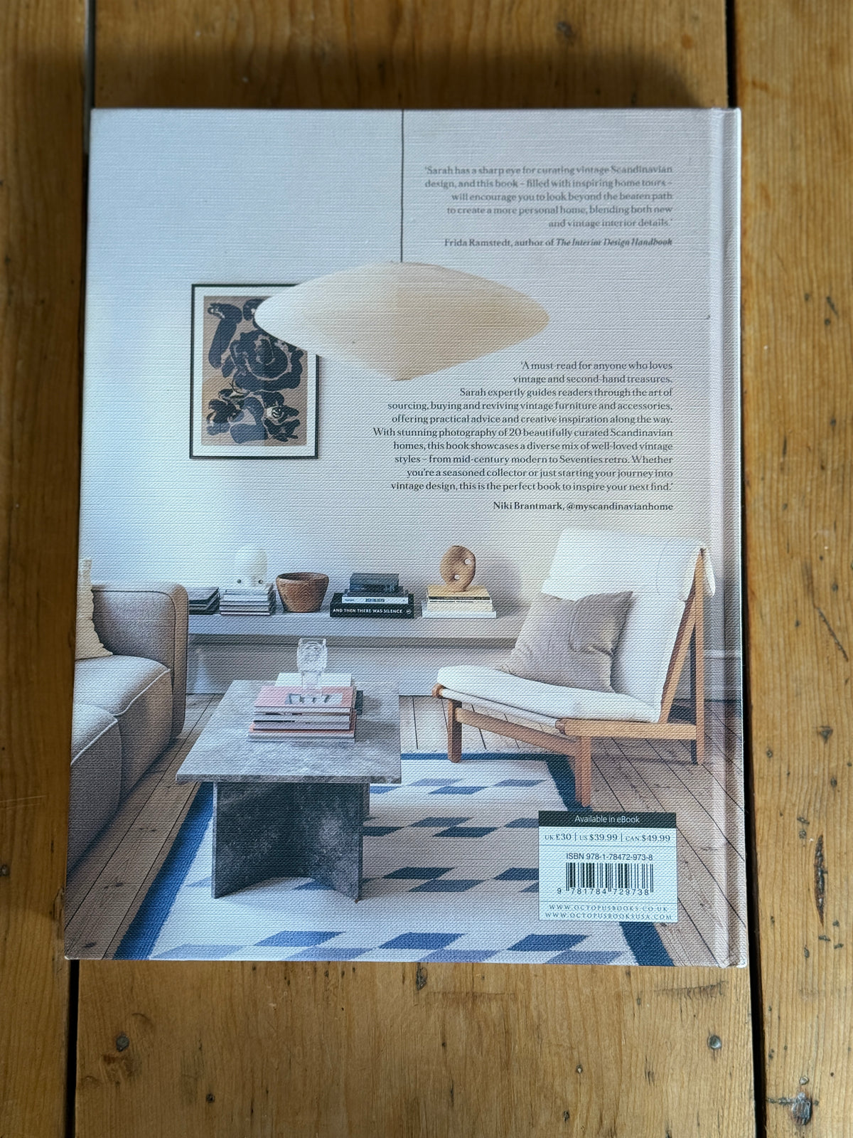 The back cover of a book titled 'Designing Interiors' by Frédéric Kervuel, featuring a styled living room interior photograph with a sofa, coffee table, rug, and chair, laid flat on a wooden surface.