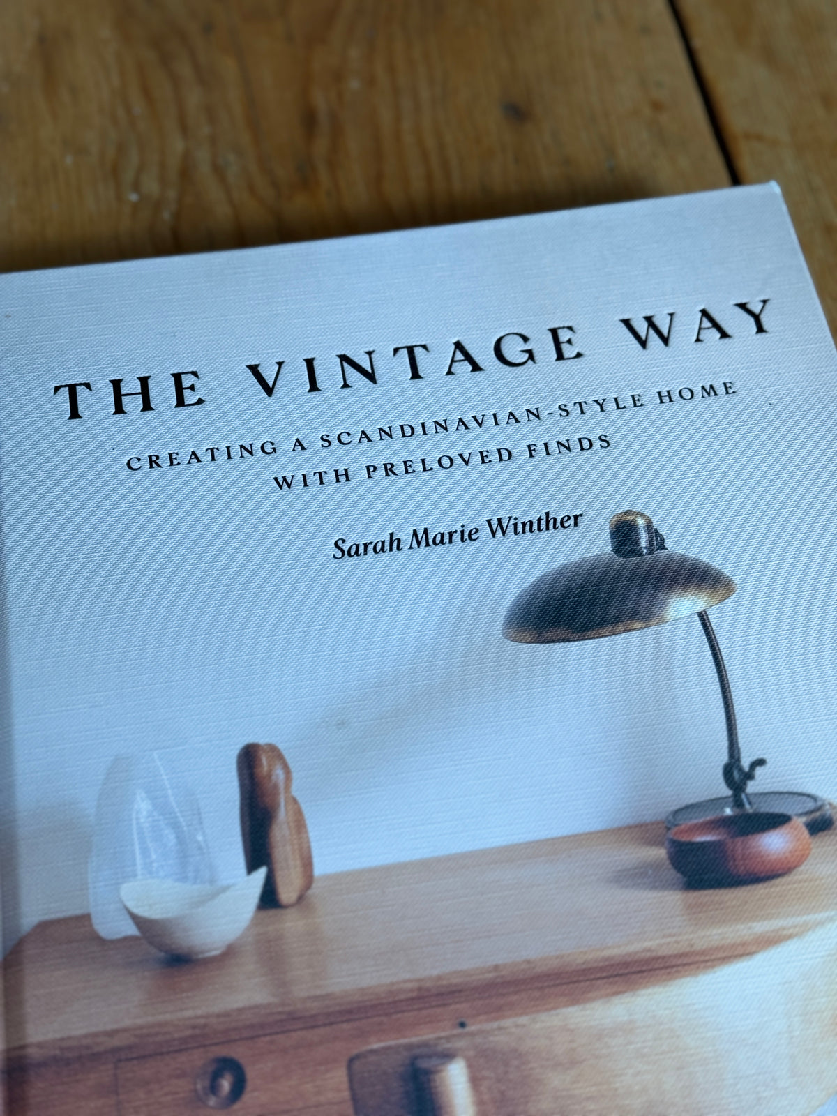 The book cover of 'The Vintage Way' by Sarah Marie Winther, styled on a wooden surface with a vintage desk lamp and small wooden objects.