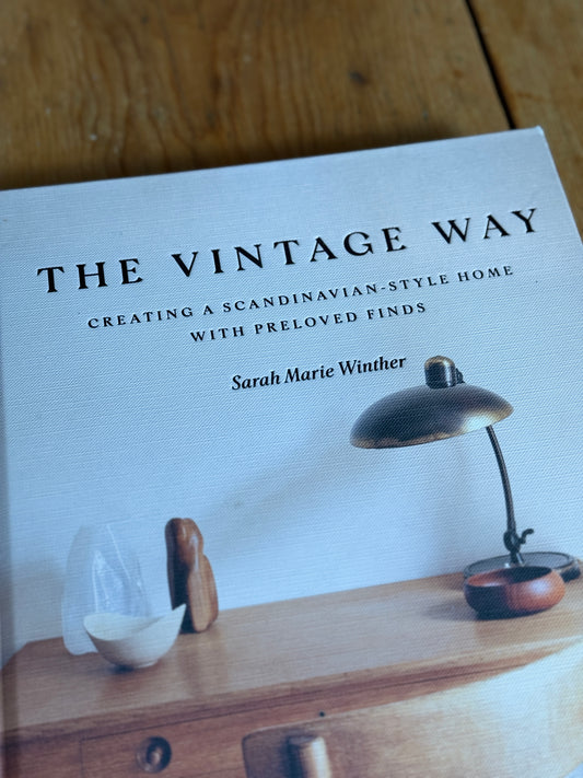 The book cover of 'The Vintage Way' by Sarah Marie Winther, styled on a wooden surface with a vintage desk lamp and small wooden objects.