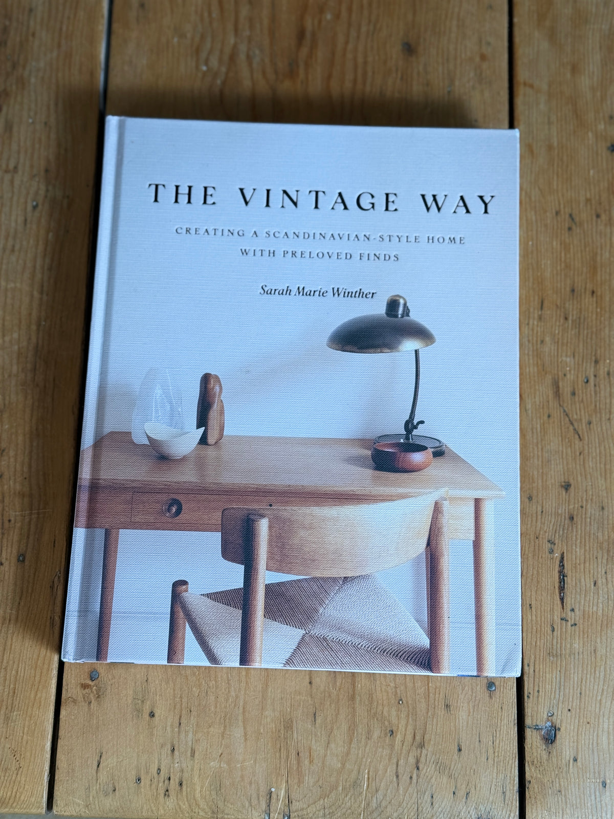 The book 'The Vintage Way' by Sarah Marie Winther, featuring a cover with a photograph of a wooden desk, chair, and lamp, displayed on a rustic wooden surface.