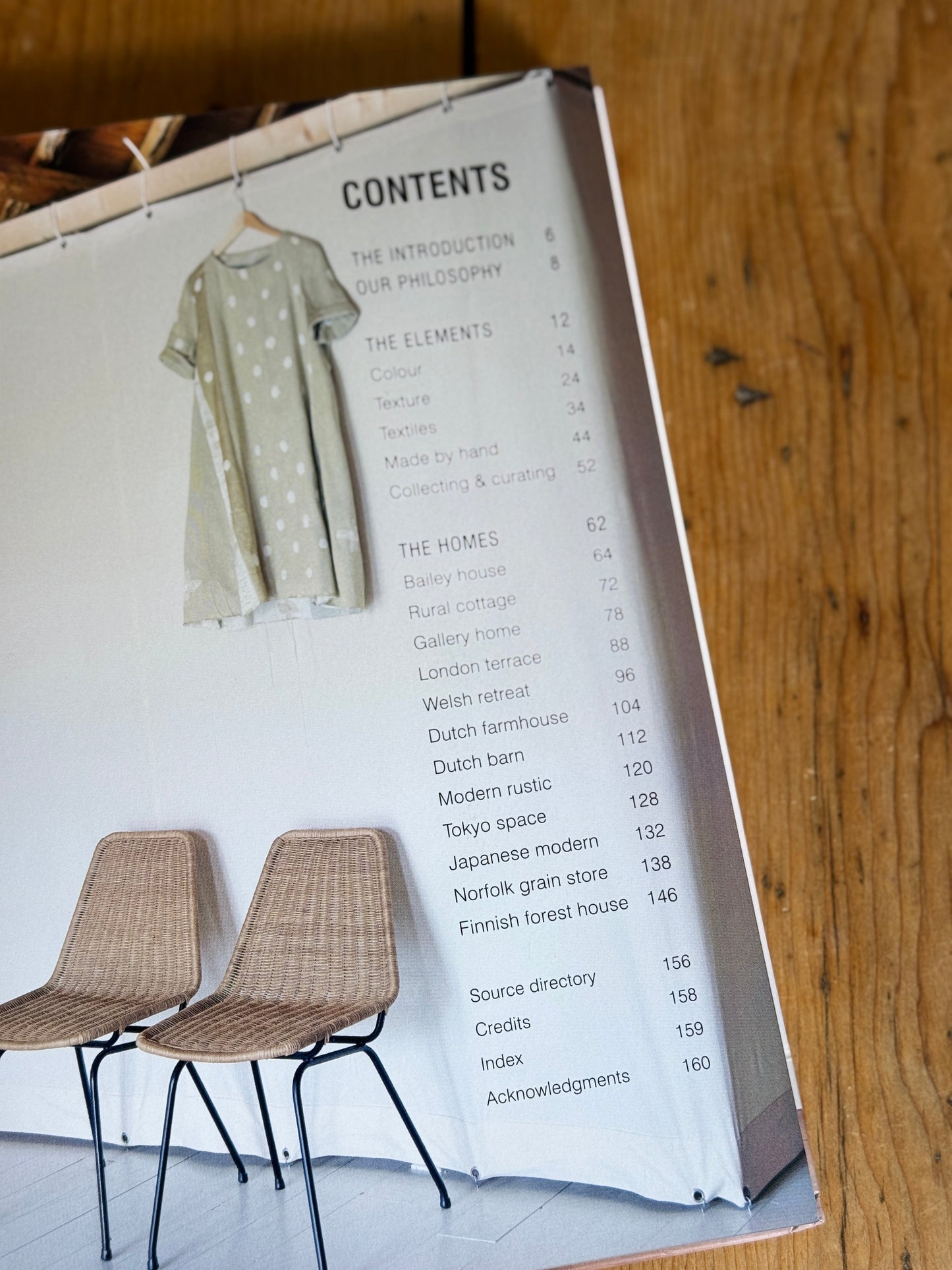 A styled flat-lay of a book's interior pages, featuring a printed table of contents, two woven chairs, and a beige polka-dot dress, all arranged on a rustic wooden tabletop.
