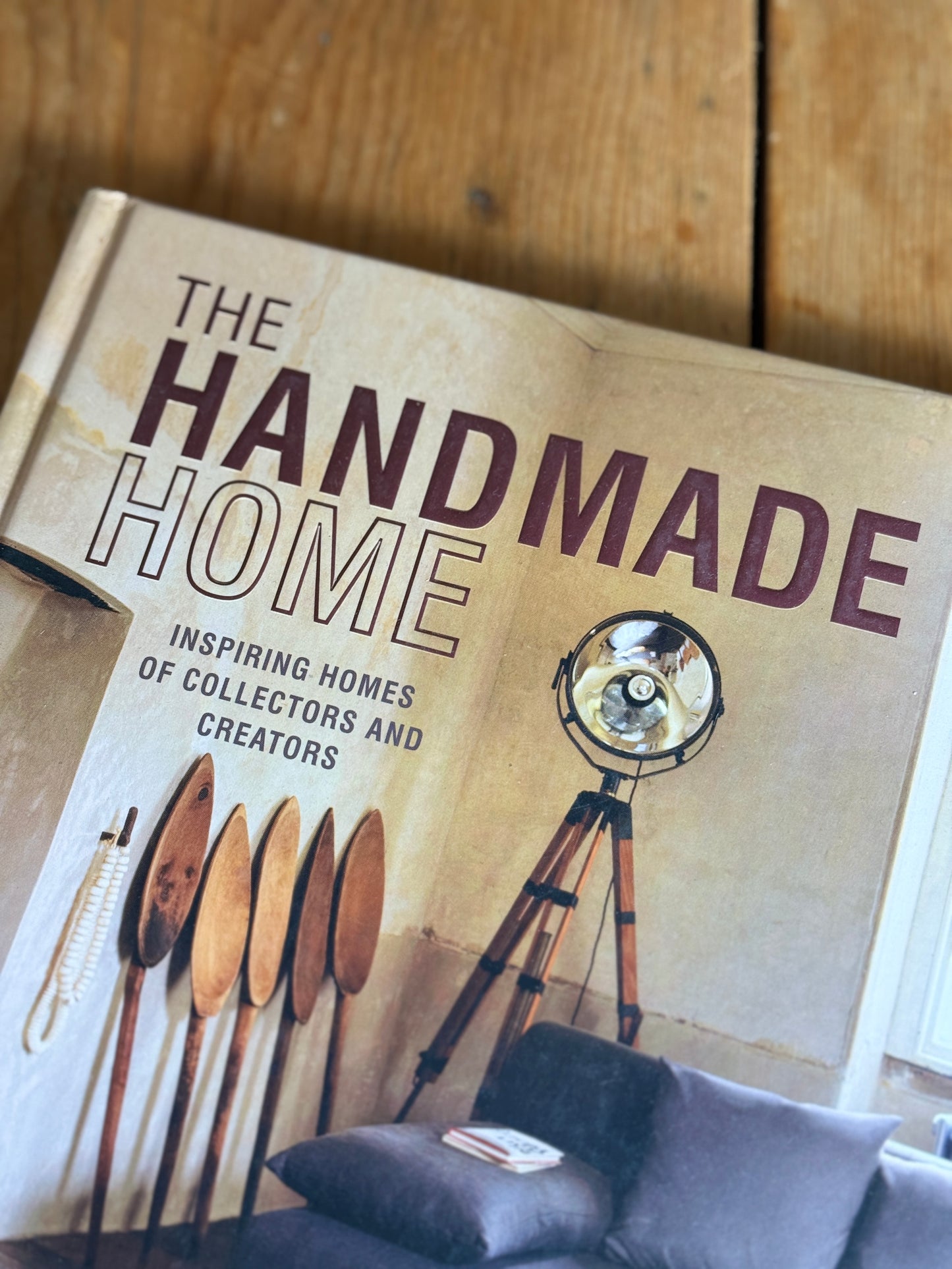 A book titled "The Handmade Home" lies open on a wooden surface, showing a beige cover with large dark text and an illustration of a tripod floor lamp. Part of a gray sofa with cushions is visible at the bottom right.