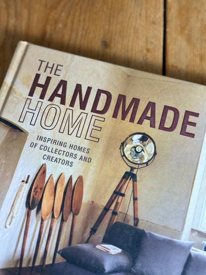 A book titled "The Handmade Home" lies open on a wooden surface, showing a beige cover with large dark text and an illustration of a tripod floor lamp. Part of a gray sofa with cushions is visible at the bottom right.