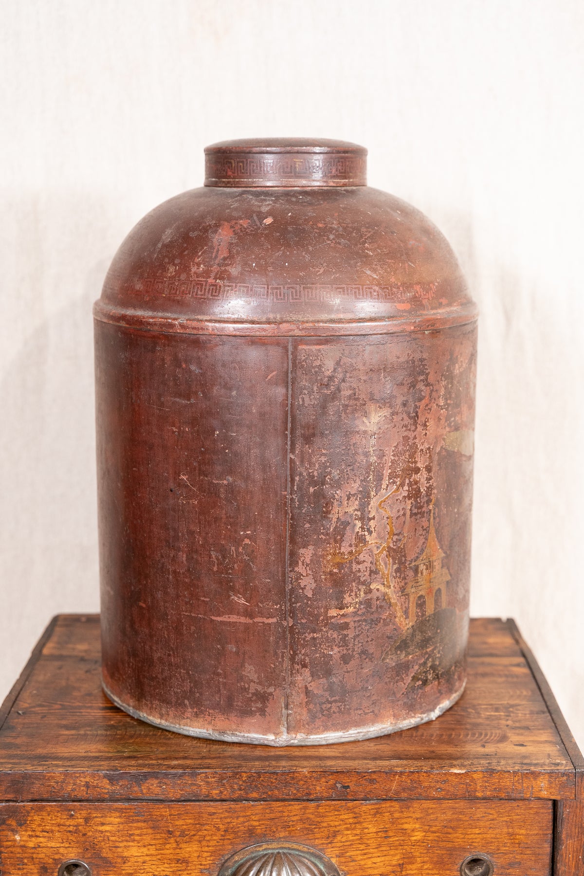 A vintage distressed brown metal canister with a domed lid, displayed on top of a wooden chest against a light off-white background.